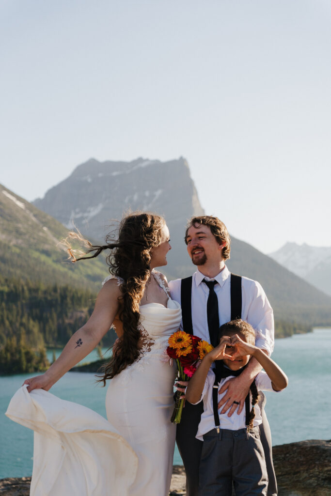 Bride and groom smile at each other while Aiden holds up a heart sign.