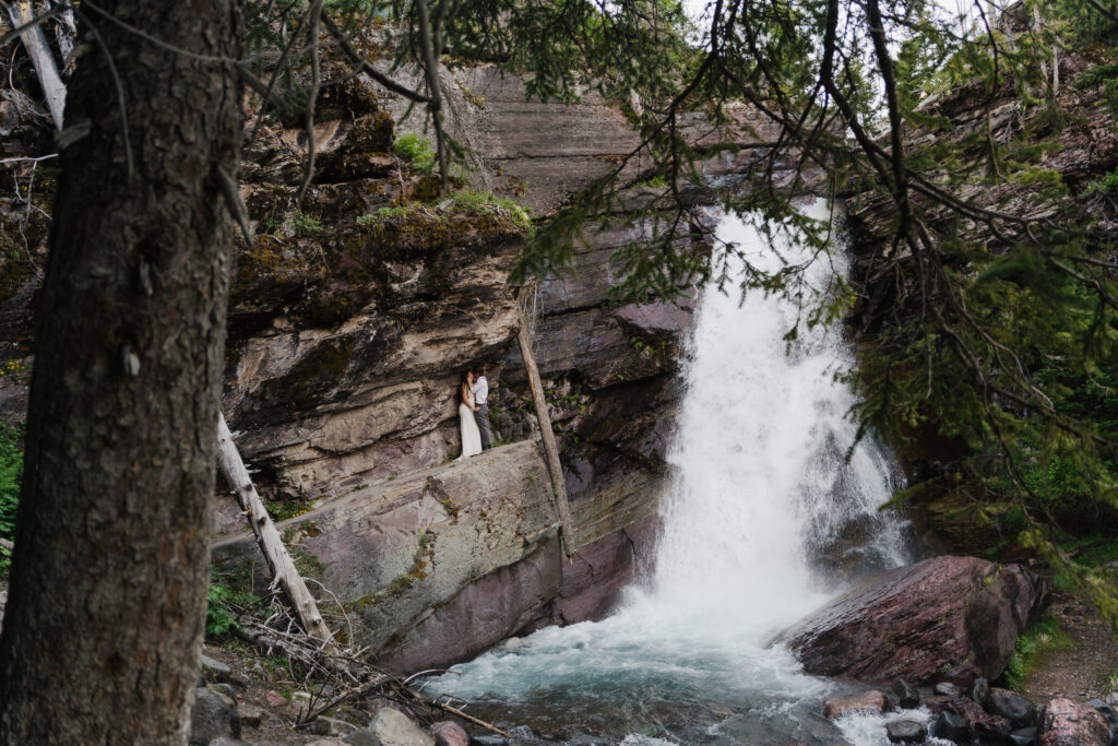 Couple standby a waterfall in Glacier National Park in their wedding outfits.