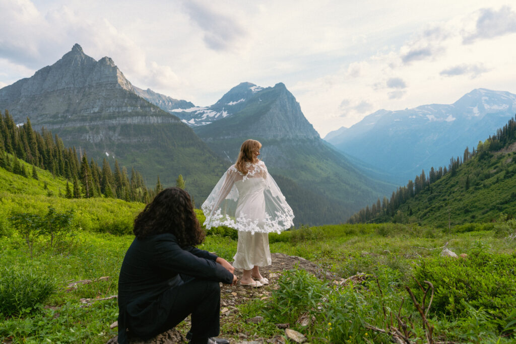 Bride spinning in bridal cape while groom smiles at her from a rock in front of a huge mountain range in Glacier National park.