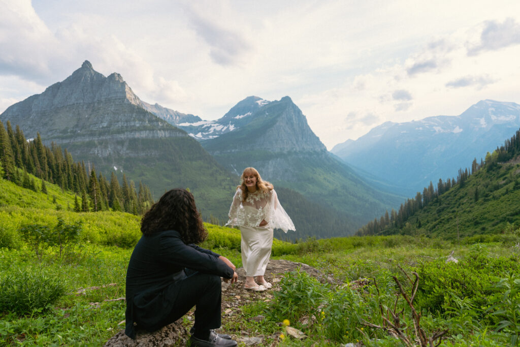 Bride spinning in bridal cape while groom smiles at her from a rock in front of a huge mountain range in Glacier National park.