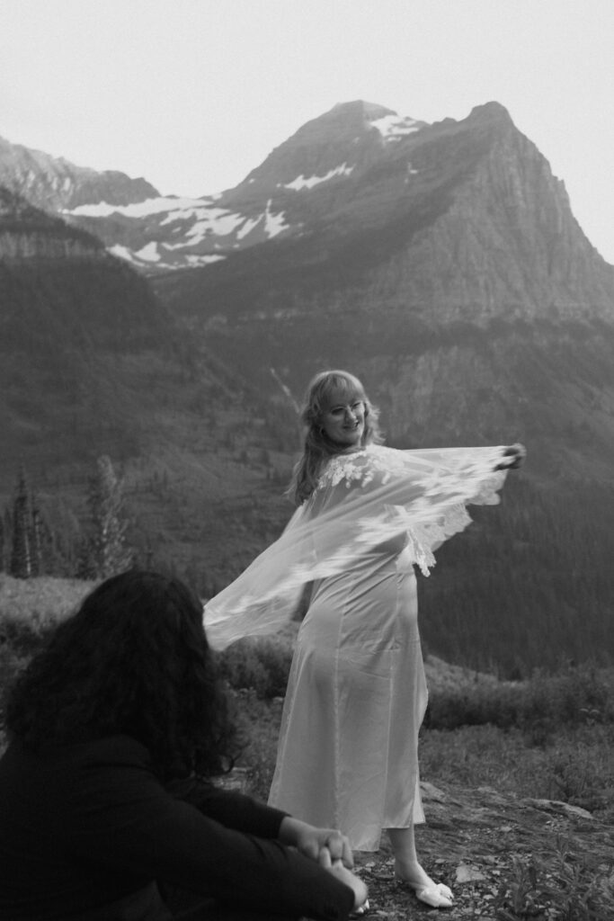 Bride spinning in bridal cape while groom smiles at her from a rock in front of a huge mountain range in Glacier National park.
