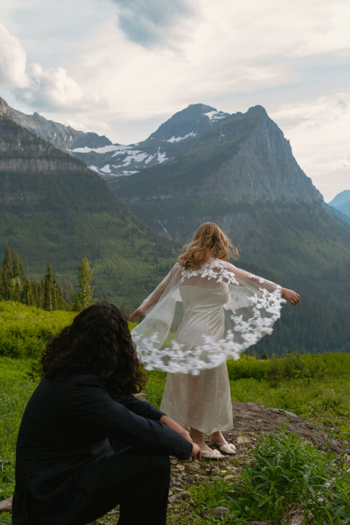Bride spinning in bridal cape while groom smiles at her from a rock in front of a huge mountain range in Glacier National park.