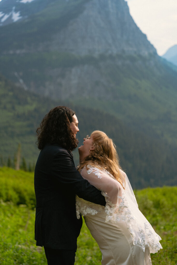 Bride and groom smiling at each other on a rock in front of a huge mountain range in Glacier National park.