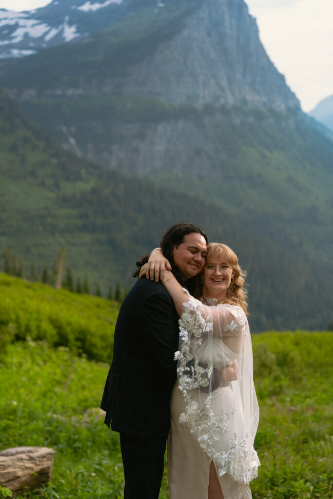 Bride and groom smiling at each other on a rock in front of a huge mountain range in Glacier National park.