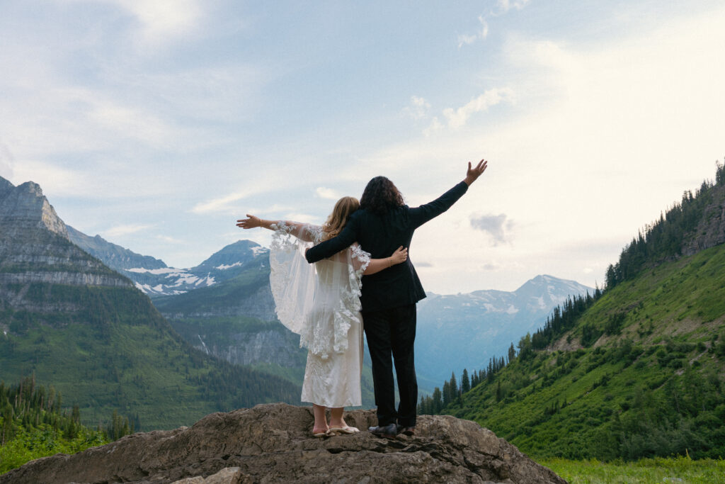 Bride and groom with arms raised looking at the mountain range in Glacier National Park.