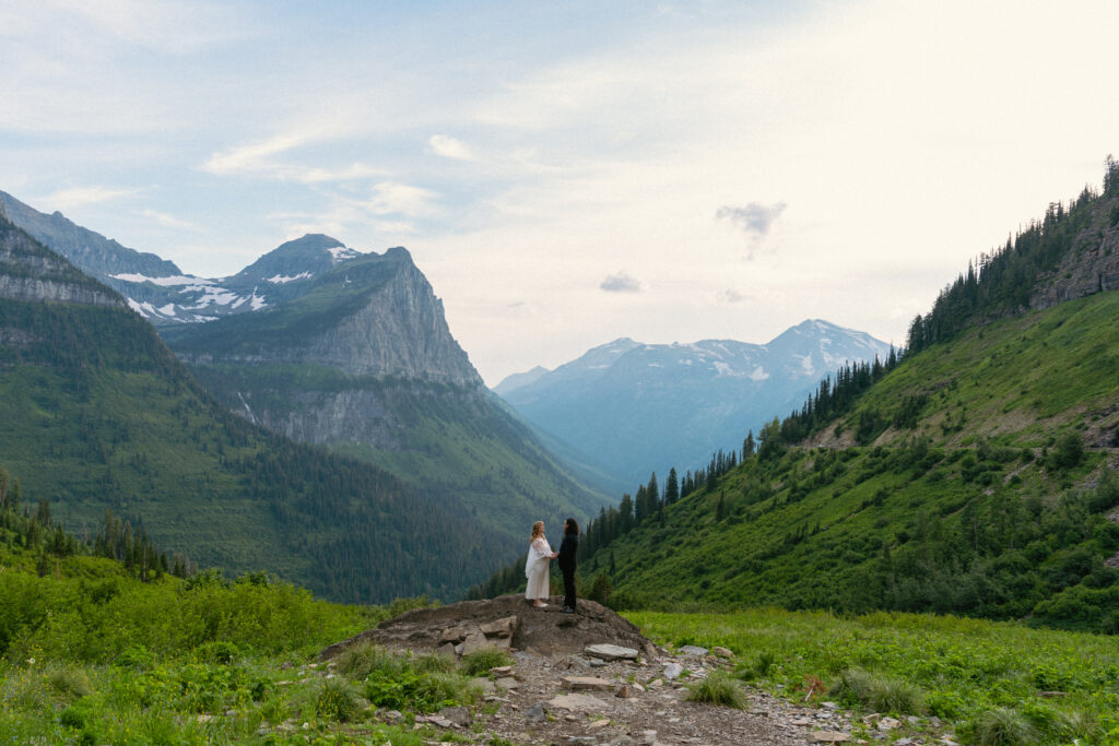 Bride and groom smiling at each other on a rock in front of a huge mountain range in Glacier National park.