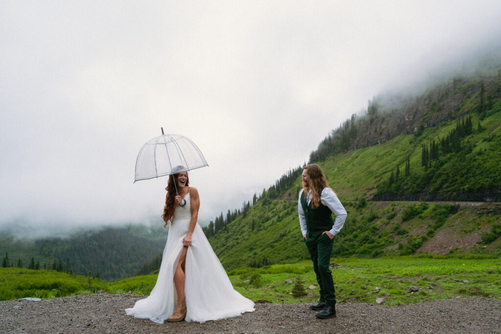 Bride laughing under an umbrella while looking at groom at a foggy spot on Going to the Sun Road at Glacier National Park