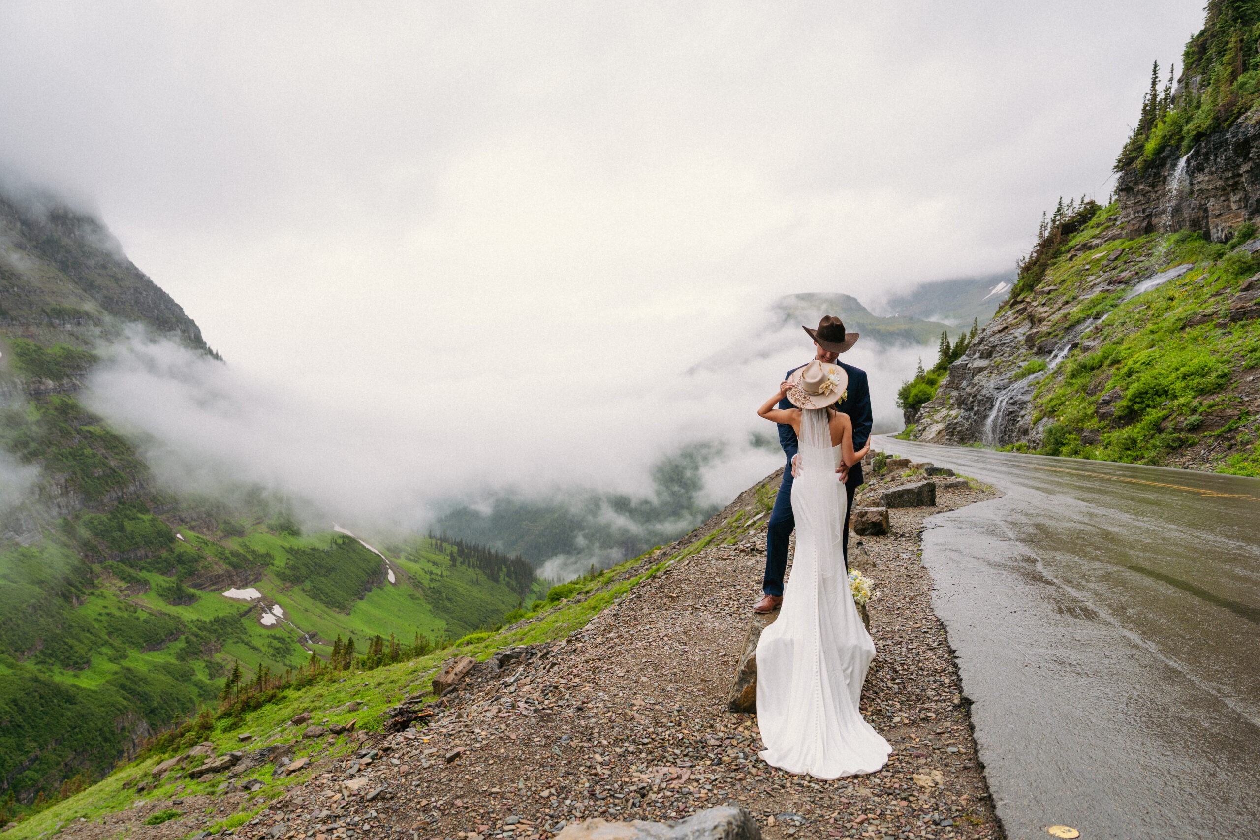 Couple stand on a rock in a cloudy Glacier National Park. The bride and groom are dress in cowboy hats.