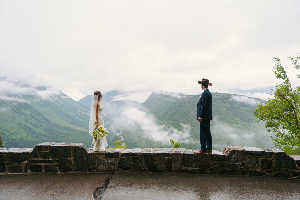 Couple stand on wall looking at river after elopement ceremony on Lake McDonald beach in Glacier National Park.